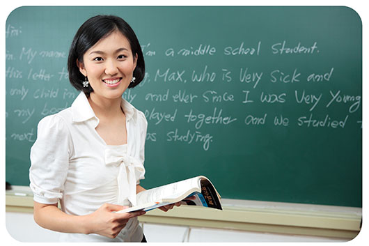 Female Asian teacher holds an open lesson plan book standing in front of a green chalkboard