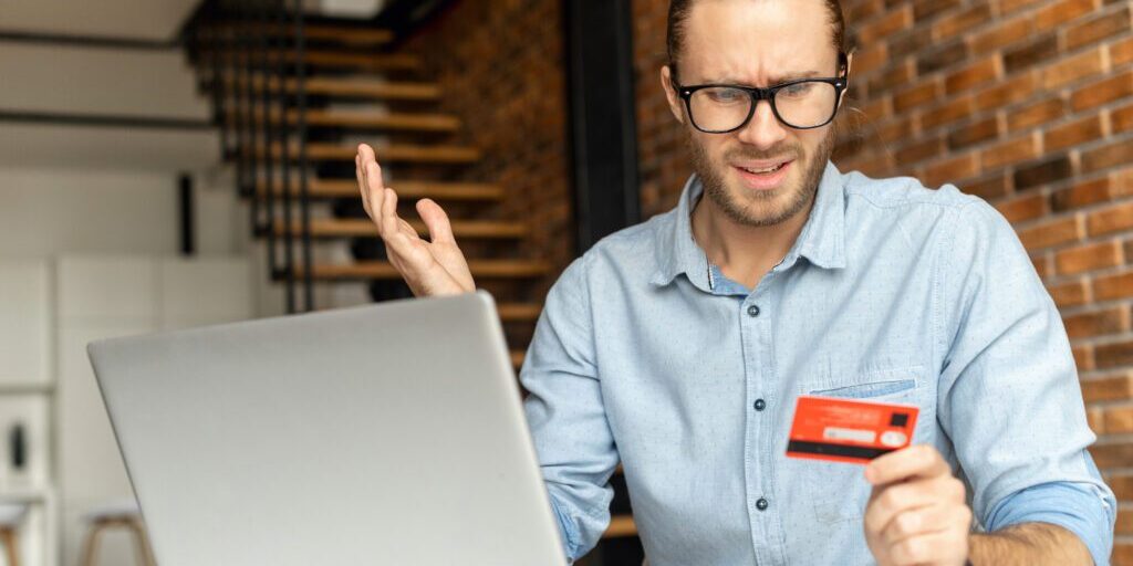 A puzzled guy sits at the desk with a laptop, holds credit card in hand and looks at it with incomprehension, a man could not complete the transaction online, not enough money on the card